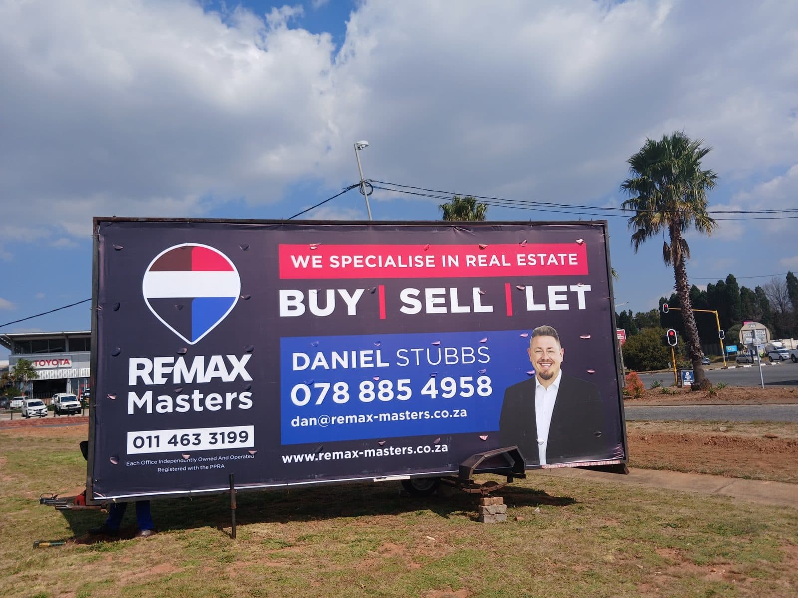 RE/MAX Masters billboard on a Simply Visible trailer at an intersection near a car dealership